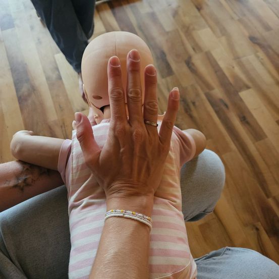 Eine Person hält eine Babypuppe und demonstriert CPR mit den Händen auf der Brust der Puppe. Die Puppe trägt ein rosa-weiß gestreiftes Shirt, und die Person hat ein Armband am Handgelenk. Eine zweite Person steht hinter ihnen auf einem Holzboden.