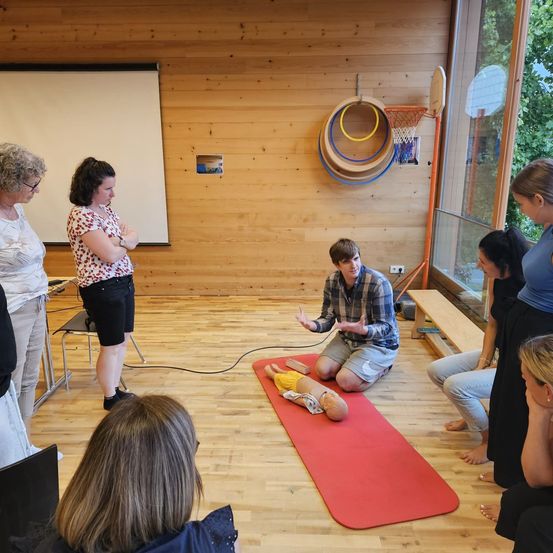 Eine Gruppe von Erwachsenen ist in einem Raum versammelt, einige sitzen auf dem Boden und andere stehen. Ein Mann kniet auf einer roten Matte und erklärt der Gruppe etwas. An der Wand befindet sich eine Tafel.