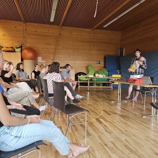 Eine Gruppe von Menschen sitzt auf Stühlen in einem Raum mit Holzböden. Ein Mann steht vor einem Tisch mit Laptop und Puppenbaby.