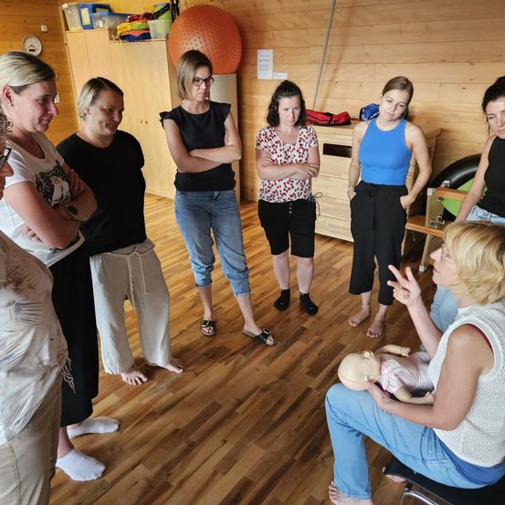 Eine Gruppe von Frauen steht im Kreis, eine hält eine Babypuppe. Eine Frau sitzt auf einem Stuhl mit einer Puppe, andere schauen zu. Holzboden, Wand und Regale sind sichtbar.