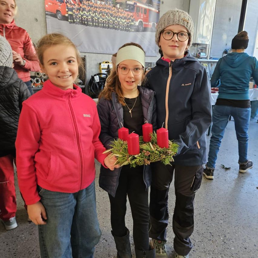 Drei Kinder stehen in einer Halle, zwei halten einen Kranz mit roten Kerzen und lächeln. Hinter ihnen steht eine Person in einer roten Jacke. Im Hintergrund ist ein Feuerwehrwagen-Poster an der Wand.