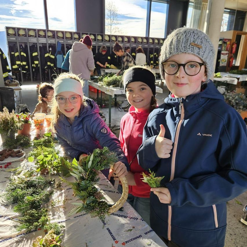 Drei Kinder lächeln und posieren in einem Raum mit einem Tisch voller Grün. Ein Mädchen hält eine Krone, und ein anderes Kind gibt einen Daumen hoch. Hinter ihnen arbeiten mehrere Personen an einem Tisch mit Pflanzen, und Feuerwehrjacken hängen an der Wand.