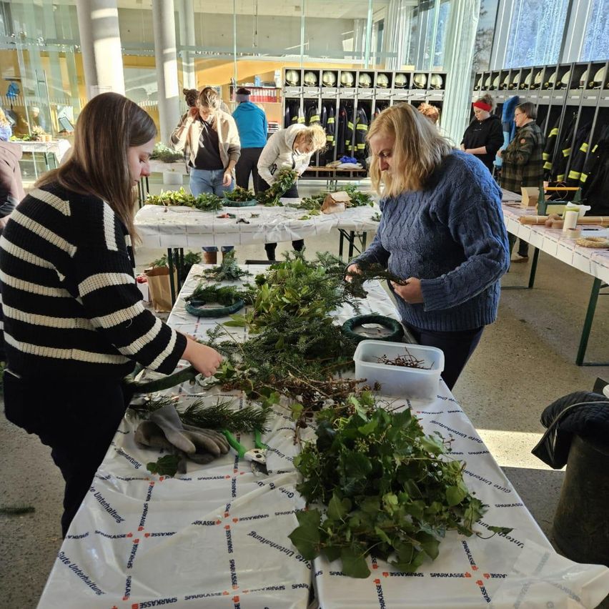 Mehrere Personen arbeiten an einem mit Grün, Handschuhen und Schnittwerkzeugen bedeckten Tisch. Ein weiterer Tisch ist mit zusätzlichem Grün bedeckt, und Feuerwehruniformen hängen im Hintergrund.