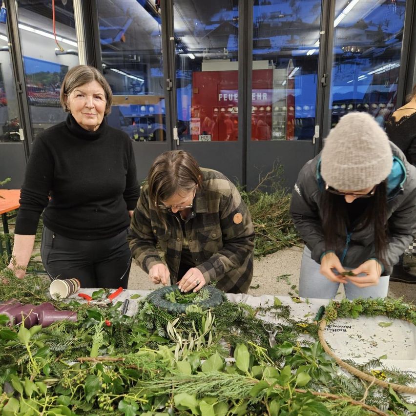 Drei Frauen stellen Kränze mit Grünzeug auf einem Tisch her, wobei eine Frau beobachtet. Hinter ihnen befindet sich eine Glastür mit einer Reflexion von Menschen.