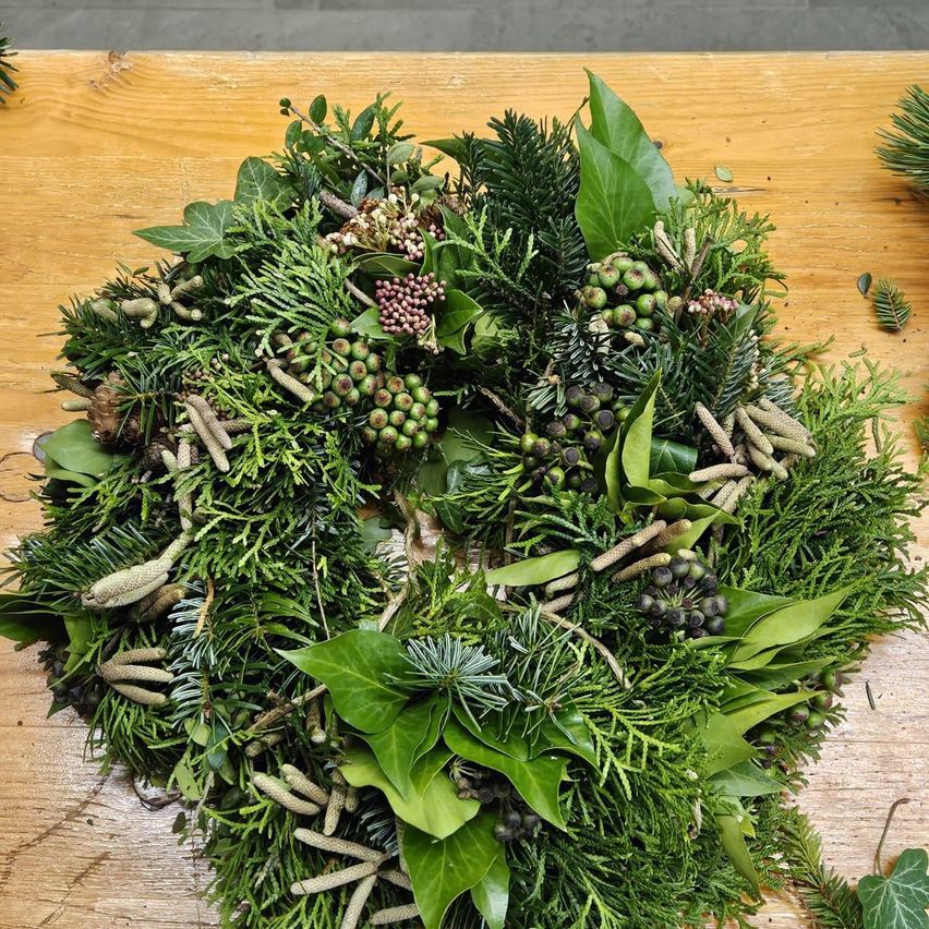 Ein Kranz aus grünen Blättern, Tannenzapfen und Beeren wird auf einer Holzoberfläche präsentiert.