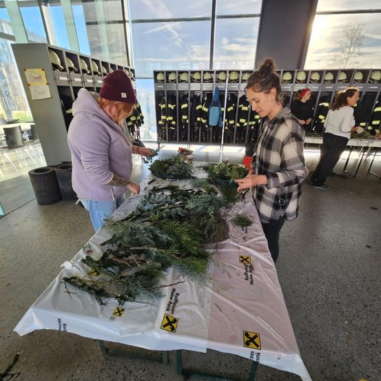 Zwei Frauen arbeiten an einem Tisch mit Tannenzweigen und Pflanzen. Sie sind von einer Feuerwache umgeben.