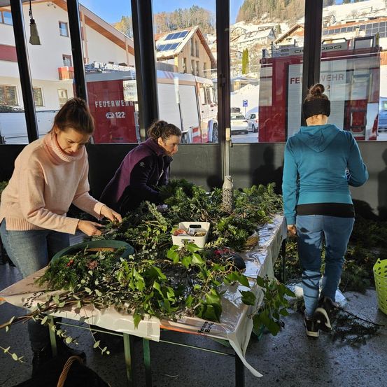 Drei Frauen arbeiten an einem mit Grünpflanzen gefüllten Tisch, möglicherweise zur Dekoration für einen Feiertag. Der Tisch ist mit Plastik bedeckt, und das Grün umfasst Stechpalme und andere Pflanzen. Die Frauen tragen Jacken und Jeans. Hinter ihnen steht ein Gebäude mit Glasfenstern.