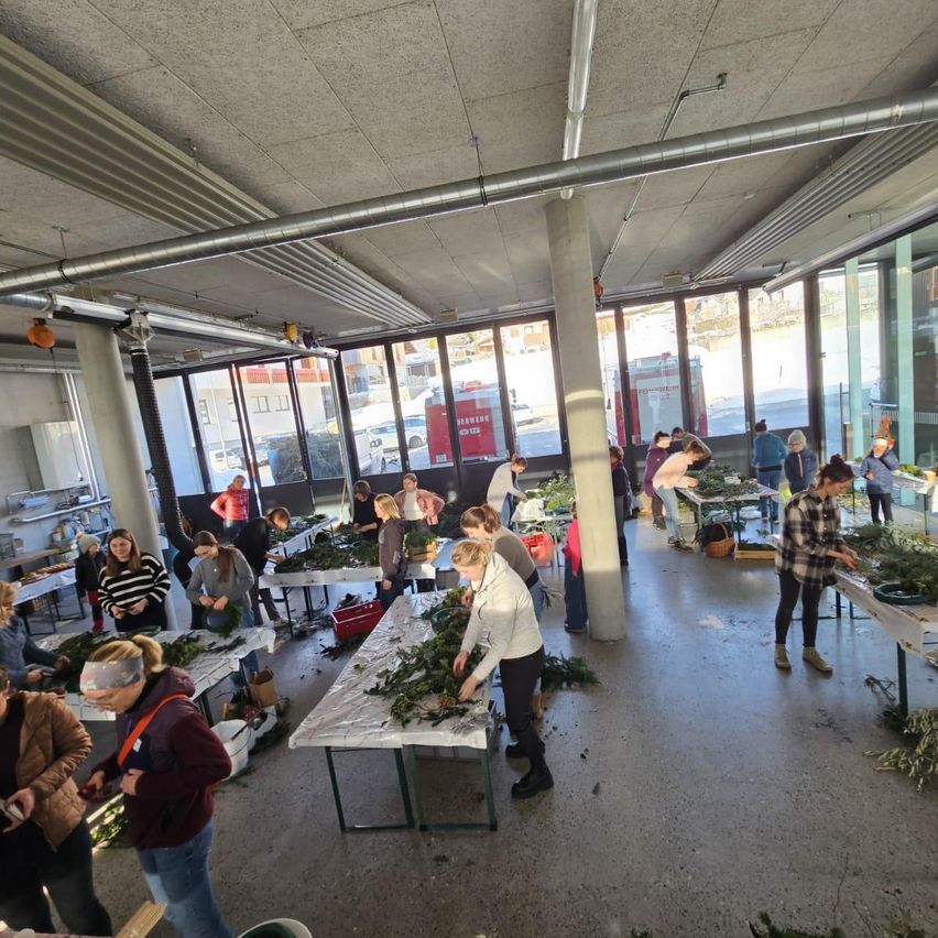 Eine große Gruppe von Menschen in einem Raum arbeitet an Tischen mit grünen Pflanzen. Der Raum hat große Fenster und ist gut beleuchtet.