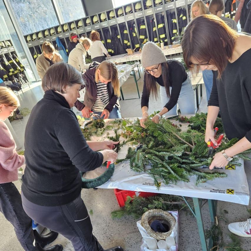 Eine Gruppe von Frauen arbeitet an einem mit Grün bedeckten Tisch, wahrscheinlich in einer Werkstatt. Sie tragen Winterkleidung und einige tragen Brillen. Sie stellen wahrscheinlich etwas mit dem Grün her.