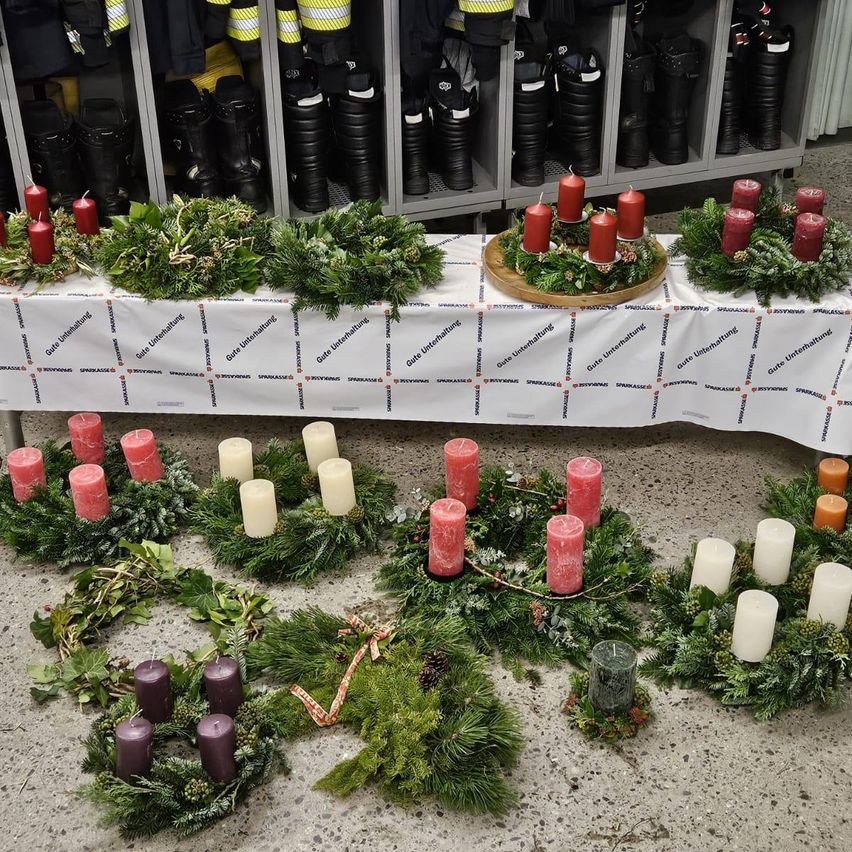 Ein Tisch ist mit einem weißen Tuch bedeckt und zeigt mehrere Kränze und Kerzen. Jeder Kranz besteht aus grünen Blättern, Tannenzapfen und roten Kerzen. Die Kerzen sind in einer Reihe angeordnet, mit einem größeren zentralen Kranz in der Mitte.