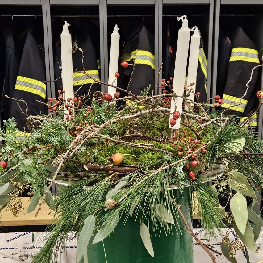 Eine festliche Dekoration mit vier weißen Kerzen, umgeben von Kiefernzweigen, Moos und roten Beeren, steht in einem grünen Eimer vor Feuerwehruniformen.