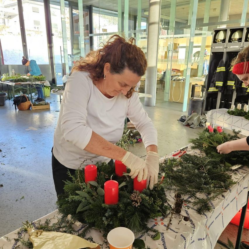 Eine erwachsene Frau mit Handschuhen arrangiert eine Weihnachtsgirlande mit roten Kerzen auf einem Tisch in einem Raum mit Glaswänden.