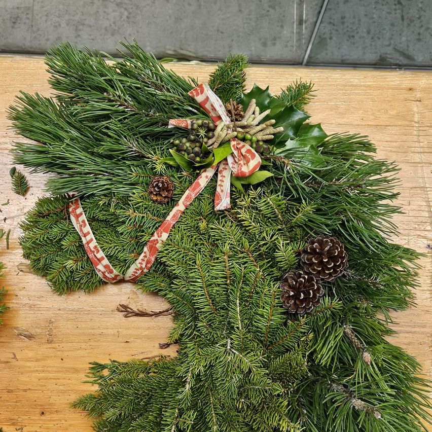 Ein Weihnachtskranz mit Tannenzweigen, Stechpalmenblättern, Tannenzapfen und einem roten Band liegt auf einer Holzoberfläche.