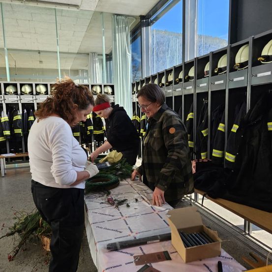 Drei Frauen arbeiten in einer Feuerwache an einem Kranz. Ein mit einem weißen Tuch bedeckter Tisch hält den Kranz. Im Hintergrund befinden sich Feuerwehrausrüstung und Helme.
