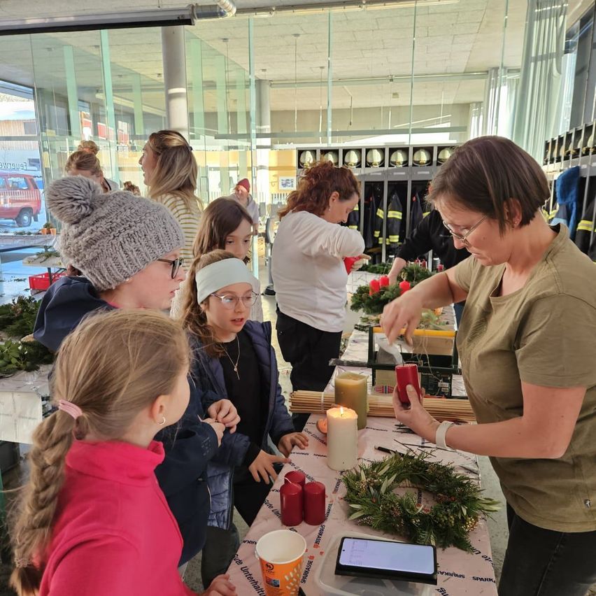 Eine Gruppe junger Mädchen lernt von einer Frau in einem braunen T-Shirt, Kränze zu machen. Sie stehen um einen Tisch mit Kerzen und Kränzen.