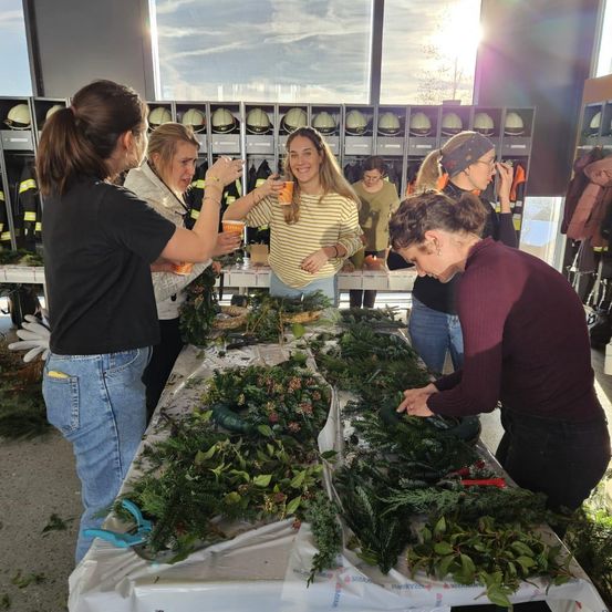 Eine Gruppe von Frauen versammelt sich um einen Tisch, der mit Grün und Ornamenten geschmückt ist, jede hält einen Becher. Hinter ihnen sind Helme an Ständern ausgestellt.
