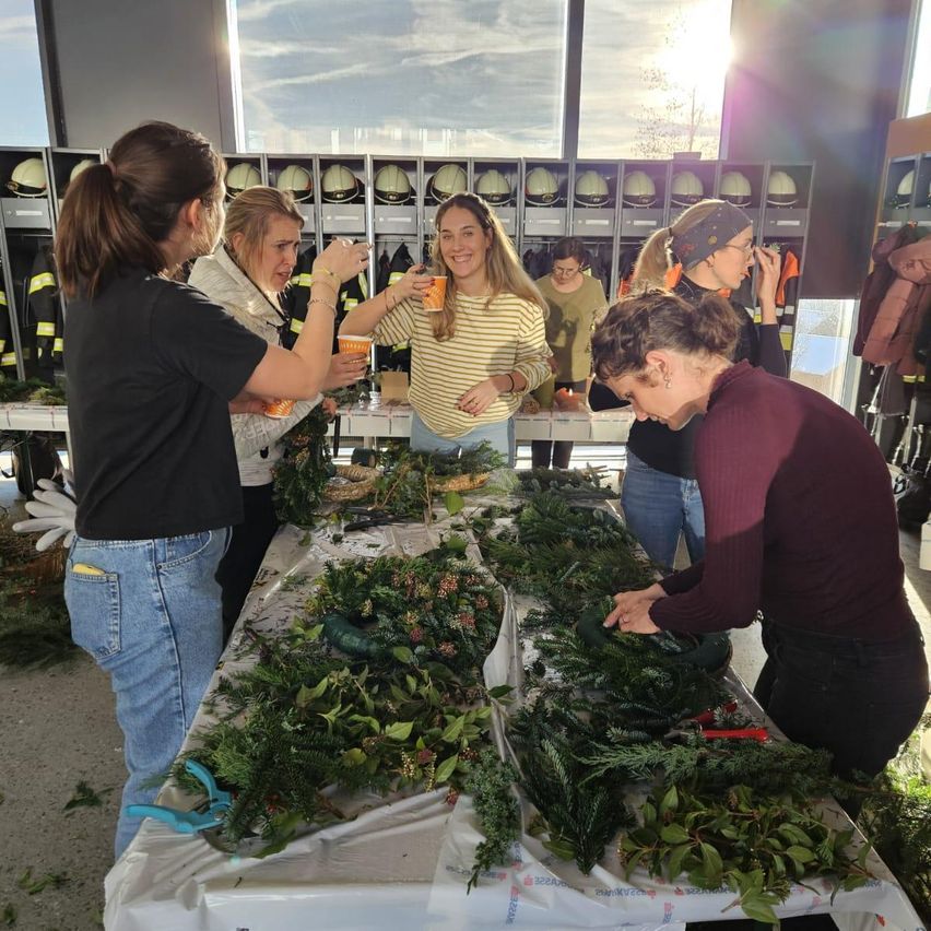 Eine Gruppe von Frauen versammelt sich um einen Tisch, der mit Grün und Ornamenten geschmückt ist, jede hält einen Becher. Hinter ihnen sind Helme an Ständern ausgestellt.