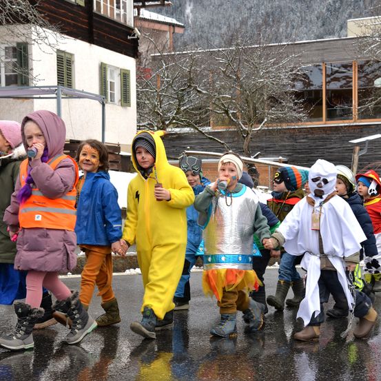Eine Gruppe von Kindern in verschiedenen Kostümen geht gemeinsam auf einer nassen Straße in einem verschneiten Gebiet. Einige tragen Gesichtsbemalung und Stiefel.