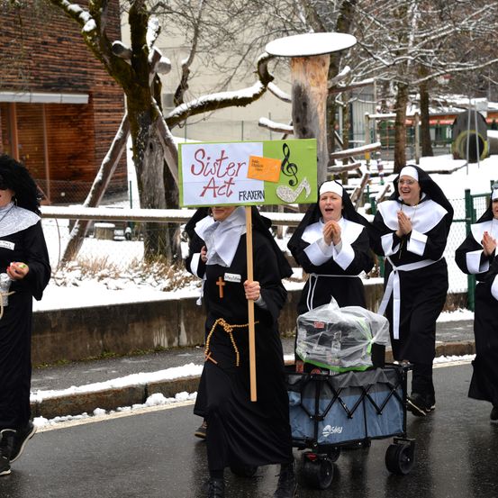 Eine Gruppe von Nonnen in Kostümen zieht in einer verschneiten Straße, hält ein Schild mit der Aufschrift 'Sister Act' und trägt einen Kinderwagen.