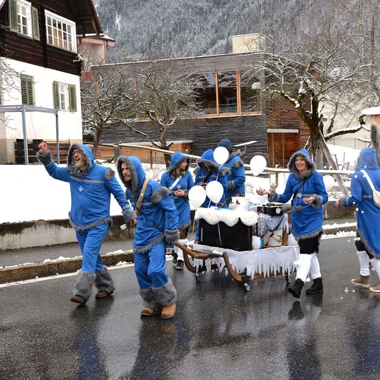 Eine Gruppe von Menschen in blauen Wintermänteln und Pelzkappe geht auf einer verschneiten Straße mit einem kleinen Wagen vor ihnen.