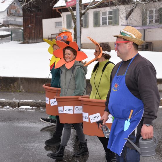 Eine Gruppe von Menschen in Kostümen geht auf einer verschneiten Straße. Sie halten Töpfe und tragen Hüte und Brillen. Der Mann trägt eine blaue Schürze.