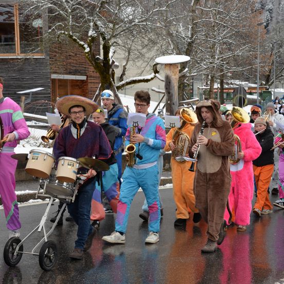 Eine Gruppe von Musikern in bunten Kostümen und Hüten marschiert auf einer nassen Straße und spielt verschiedene Instrumente wie Trommeln, Tubas und Saxophone. Dahinter stehen Gebäude mit schneebedeckten Dächern und Bäumen.