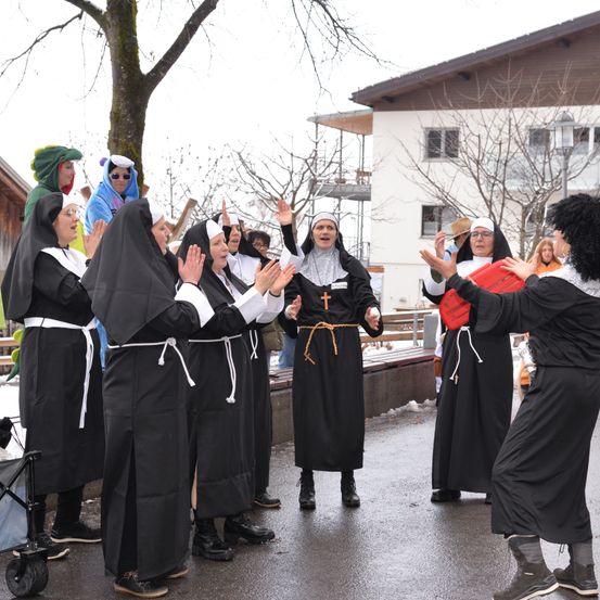 Eine Gruppe von Menschen, die als Nonnen verkleidet sind, singen und feiern im Freien, in der Nähe eines Gebäudes mit Bänken und Bäumen.