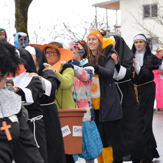 Eine Gruppe von Menschen in Nonnentracht, einige mit Hüten, stehen auf einem Außengelände. Eine Person hält einen Eimer mit der Aufschrift 'Gem'.