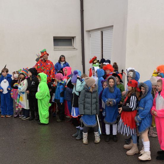 Eine Gruppe von Menschen, darunter Kinder, in verschiedenen farbenfrohen Kostümen gekleidet, steht in einer Reihe vor einem Gebäude. Ein Mann in einem Clownskostüm steht vorne.