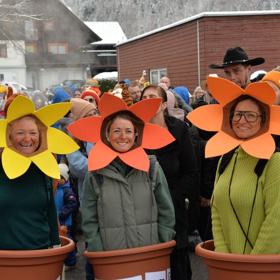 Drei Frauen mit Blumenhüten stehen in einer Menschenmenge während eines Festivals. Dahinter sind eine Menschenmenge und Gebäude zu sehen.