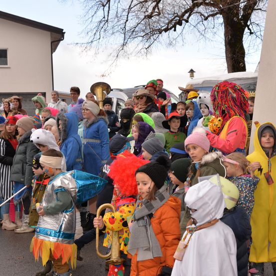 Eine Menschenmenge in farbenfrohen Kostümen, Hüten und Schuhen versammelt sich auf der Straße für einen Umzug. Einige tragen Schminke und Masken. Fahrzeuge sind in der Nähe geparkt.