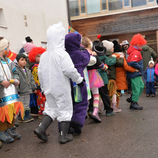 Kinder in verschiedenen bunten Kostümen sind draußen versammelt, einige mit Gesichtsbemalung. Ein Gebäude mit dem Namen Jakob Summers ist im Hintergrund.