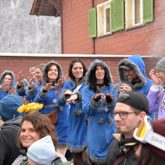 Eine Gruppe von Menschen in blauen Kostümen mit Pelzbesatz steht vor einem Gebäude mit grünen Fensterläden. Einige tragen Brillen und Mützen. Sie scheinen an einer festlichen Veranstaltung teilzunehmen oder aufzutreten.