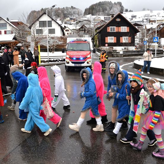 In einem verschneiten Dorf überquert eine Gruppe von Kindern in bunten Kostümen die Straße, während ein Feuerwehrauto geparkt ist. Leute stehen herum, und eine Frau macht ein Foto.