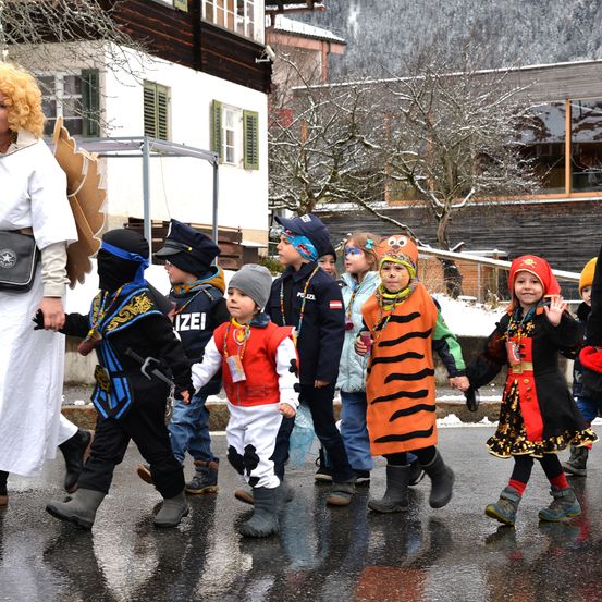 Eine Gruppe von Kindern in Kostümen geht auf einer nassen Straße, angeführt von einer Frau, mit Gebäuden im Hintergrund.