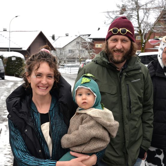 Eine Familie steht auf einer verschneiten Straße und lächelt. Der Mann trägt eine grüne Jacke und einen roten Hut mit einer Sonnenbrille. Die Frau hält ein Baby in einer grünen Kapuze. Eine weitere Person in einem Kuhkostüm steht hinter ihnen.