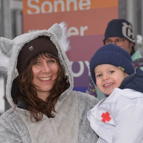 Eine Frau in einem grauen Wolfskostüm und ein Kind in einem weißen Mantel mit rotem Kreuz lächeln für die Kamera. Dahinter ist ein Schild mit orangen und roten Farben zu sehen.