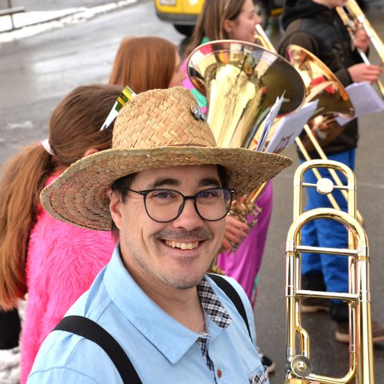 Ein Mann in einem Strohhut und mit Brille lächelt für die Kamera, mit einem Blasorchester im Hintergrund. Die Bandmitglieder halten Blechblasinstrumente.