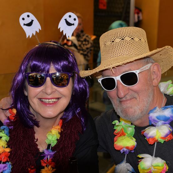 Ein Mann und eine Frau lächeln für ein Foto, beide tragen Sonnenbrillen und bunte Girlanden. Die Frau hat lila Haare mit Geister-Haarbändern und der Mann einen Strohhut. Sie scheinen auf einer Veranstaltung zu sein.