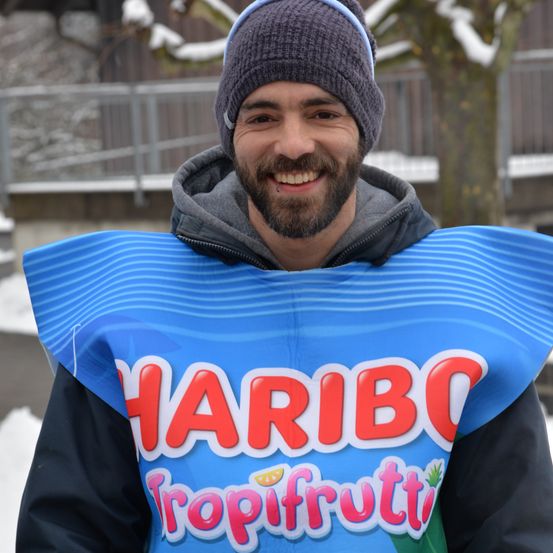Ein lächelnder Mann in einer grauen Kapuzenjacke und einer grauen Mütze steht im Schnee. Er trägt ein blaues Kostüm mit dem Wort 'HARIBO' und 'Tropifruitti' darauf geschrieben.