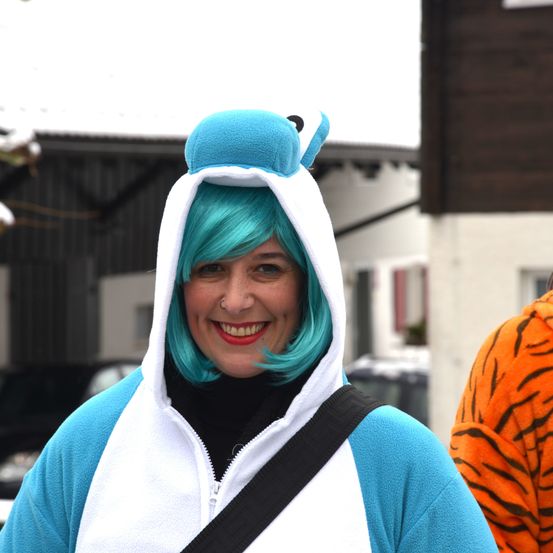 Eine Frau in einem blau-weißen Kostüm mit grünen Haaren lächelt und trägt einen schwarzen Riemen. Ein Tigerkostüm ist hinter ihr sichtbar.