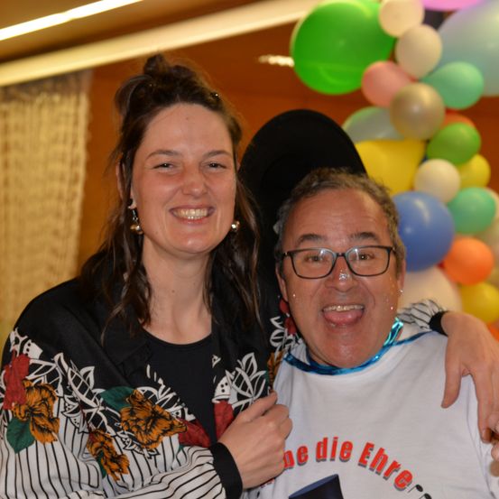 Eine lächelnde Frau mit einem schwarzen Hut und ein Mann mit Brille posieren für ein Foto vor einem bunten Ballonhintergrund.