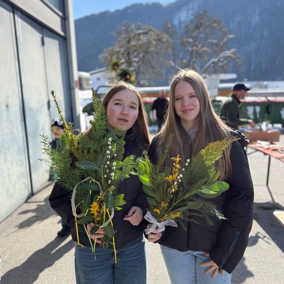 Zwei Mädchen stehen an einem sonnigen Tag draußen und halten jeweils einen Blumenstrauß mit gelben und grünen Blättern. Sie tragen schwarze Jacken und Jeans. Im Hintergrund befinden sich Berge, Bäume und ein Gebäude.