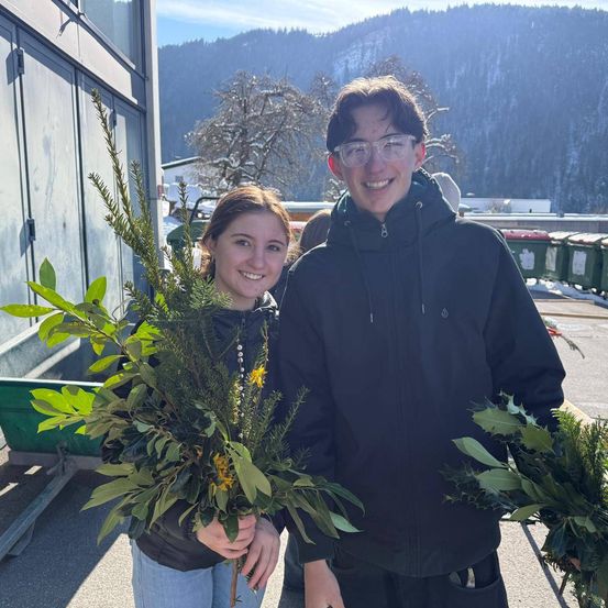 Ein Mann und eine Frau posieren draußen mit Girlanden in den Händen. Sie stehen vor einem Gebäude mit einem verschneiten Berg im Hintergrund.