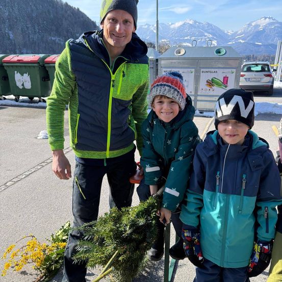 Ein Mann und zwei Kinder stehen neben einem Weihnachtsbaum in einem verschneiten Gebiet. Der Mann lächelt und hält den Baum, während die Kinder neben ihm lächeln. Dahinter befinden sich Mülleimer und eine Bergkette.