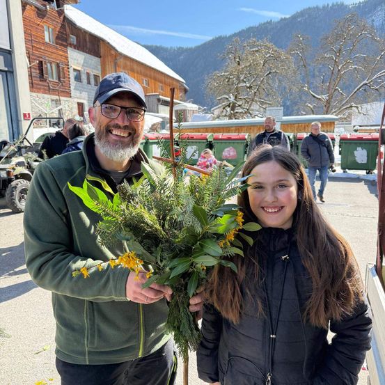 Ein Mann und ein Mädchen stehen in einer verschneiten Landschaft und halten einen Blumenstrauß. Hinter ihnen sind Gebäude, Berge und Menschen, die gehen.