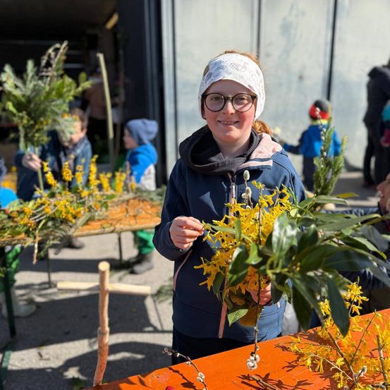 Ein Mädchen mit Brille und blauer Jacke hält gelbe Blumen in einer Außenumgebung, mit anderen Kindern und Erwachsenen in der Umgebung.