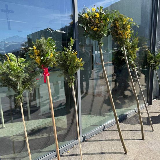 Vier Blumenarrangements mit je einem roten Band werden vor einem Gebäude mit Glaswänden ausgestellt. Spiegelungen von Menschen und einem Kreuz sind im Glas zu sehen.