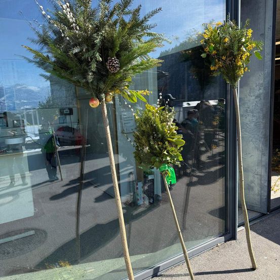 Drei dekorative baumartige Anordnungen mit Blättern und Früchten stehen vor einer Glaswand. Die Szene beinhaltet Spiegelungen von Menschen und Gegenständen im Inneren.