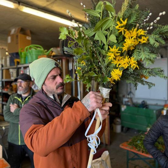 Ein Mann in einer grünen Mütze und einem orangefarbenen Mantel hält einen großen Blumenstrauß in einem Raum mit Regalen und Lagerboxen. Ein weiterer Mann in einem grünen Mantel steht hinter ihm.